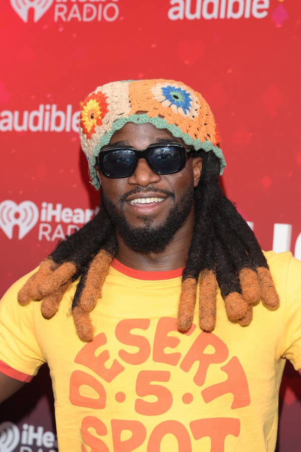 Man in crochet hat and sunglasses on a red carpet, wearing a yellow &quot;Desert 5 Spot&quot; shirt