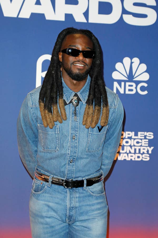 I don&#x27;t know who this is. Person on red carpet wearing denim outfit, sunglasses, and a decorative belt. NBC and &quot;People&#x27;s Choice Country Awards&quot; logos are visible