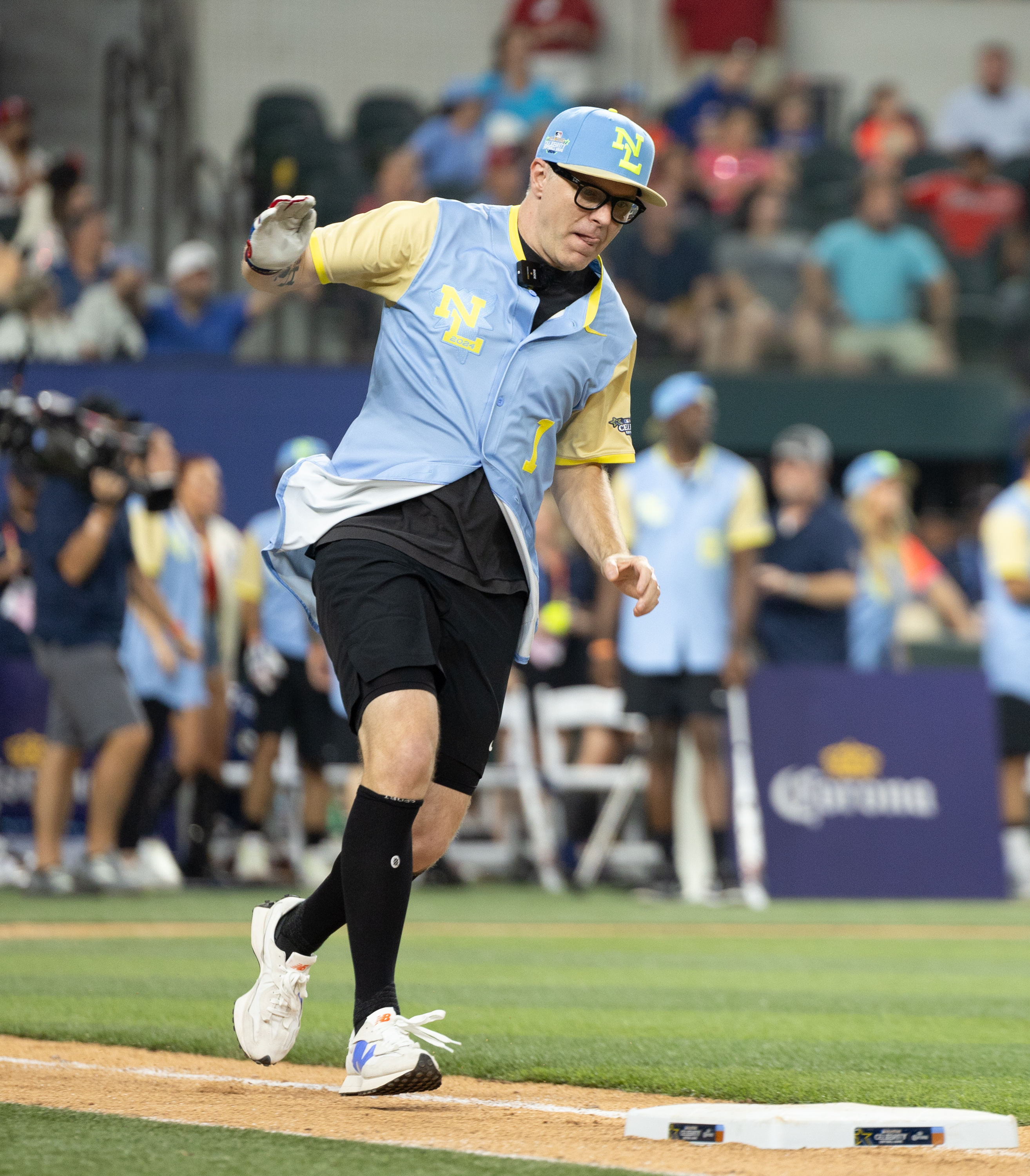 Bobby Bones running a base at a celebrity baseball game, wearing a jersey and cap. Crowd and camera operators in the background