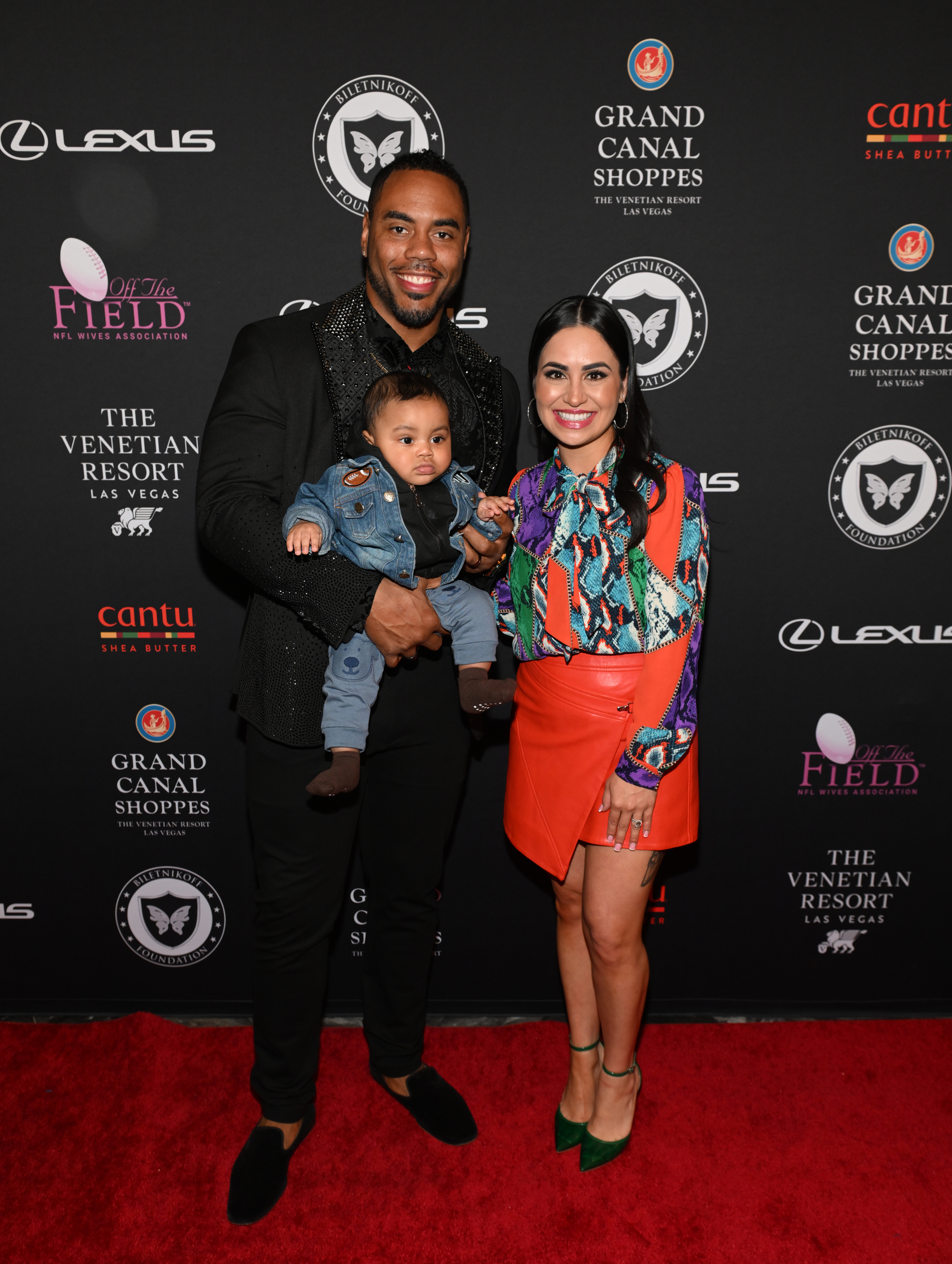 Rashad Jennings holds a baby and stands beside Destiny Jennings on a red carpet. The woman wears a colorful blouse and an orange skirt.