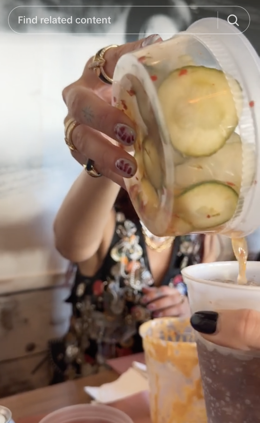 A person pours cucumber slices into a cup. They have nail art and wear multiple rings and a floral-patterned top