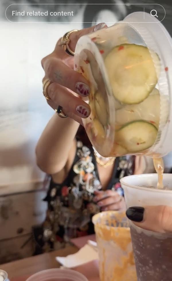 A person pours cucumber slices into a cup. They have nail art and wear multiple rings and a floral-patterned top