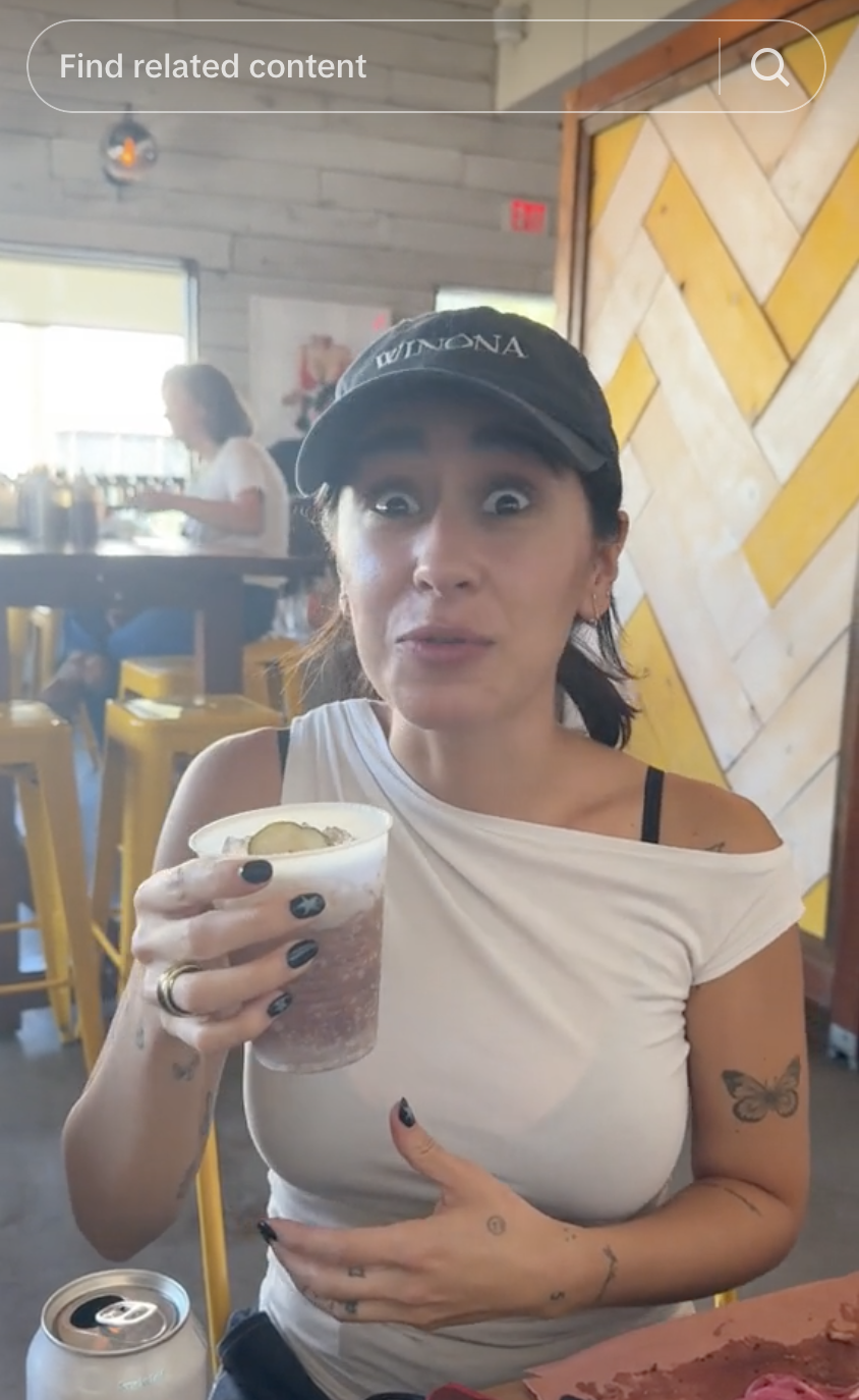 Woman in a casual eatery, wearing a black cap and white off-shoulder top, holding a drink with a surprised expression