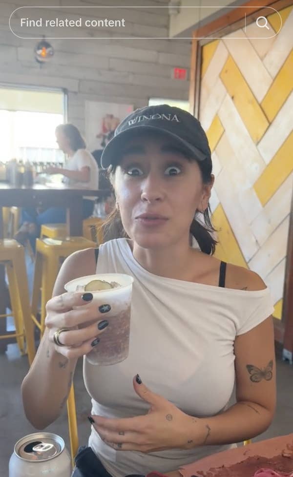 Woman in a casual eatery, wearing a black cap and white off-shoulder top, holding a drink with a surprised expression