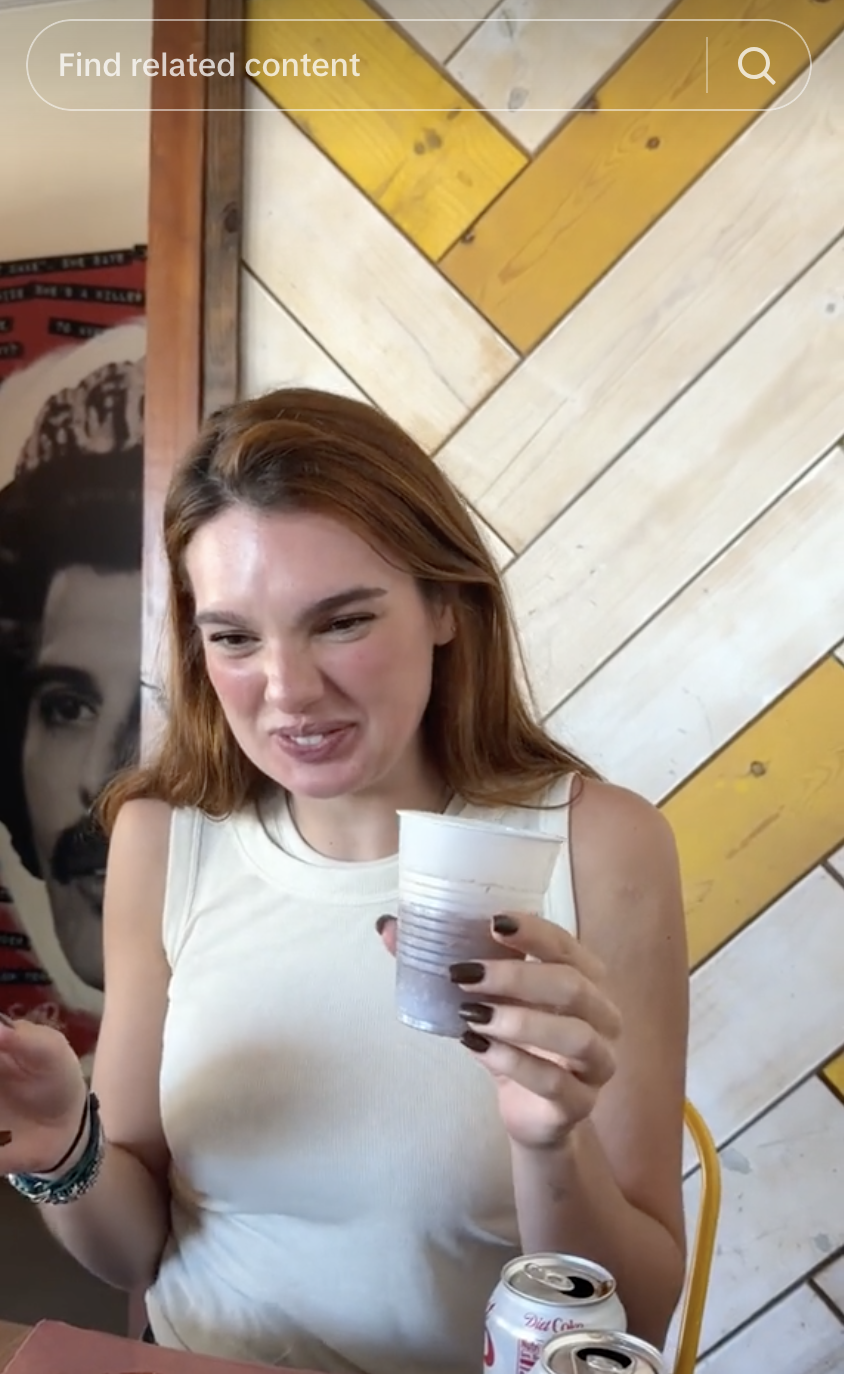 A woman holding a cup and smiling in a casual setting with a decorative wall behind her