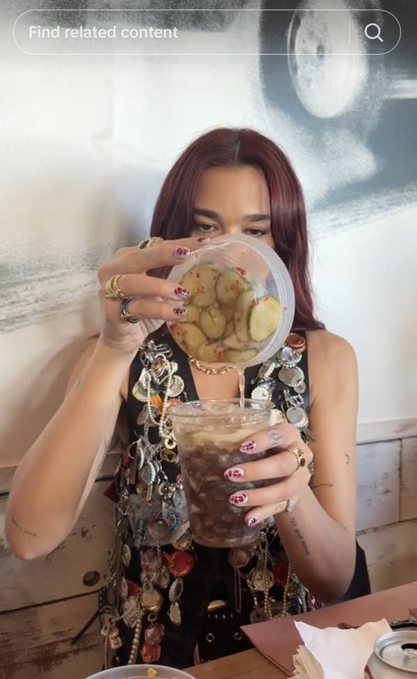 A person with long hair holds a lid with pickles above a drink in a casual setting