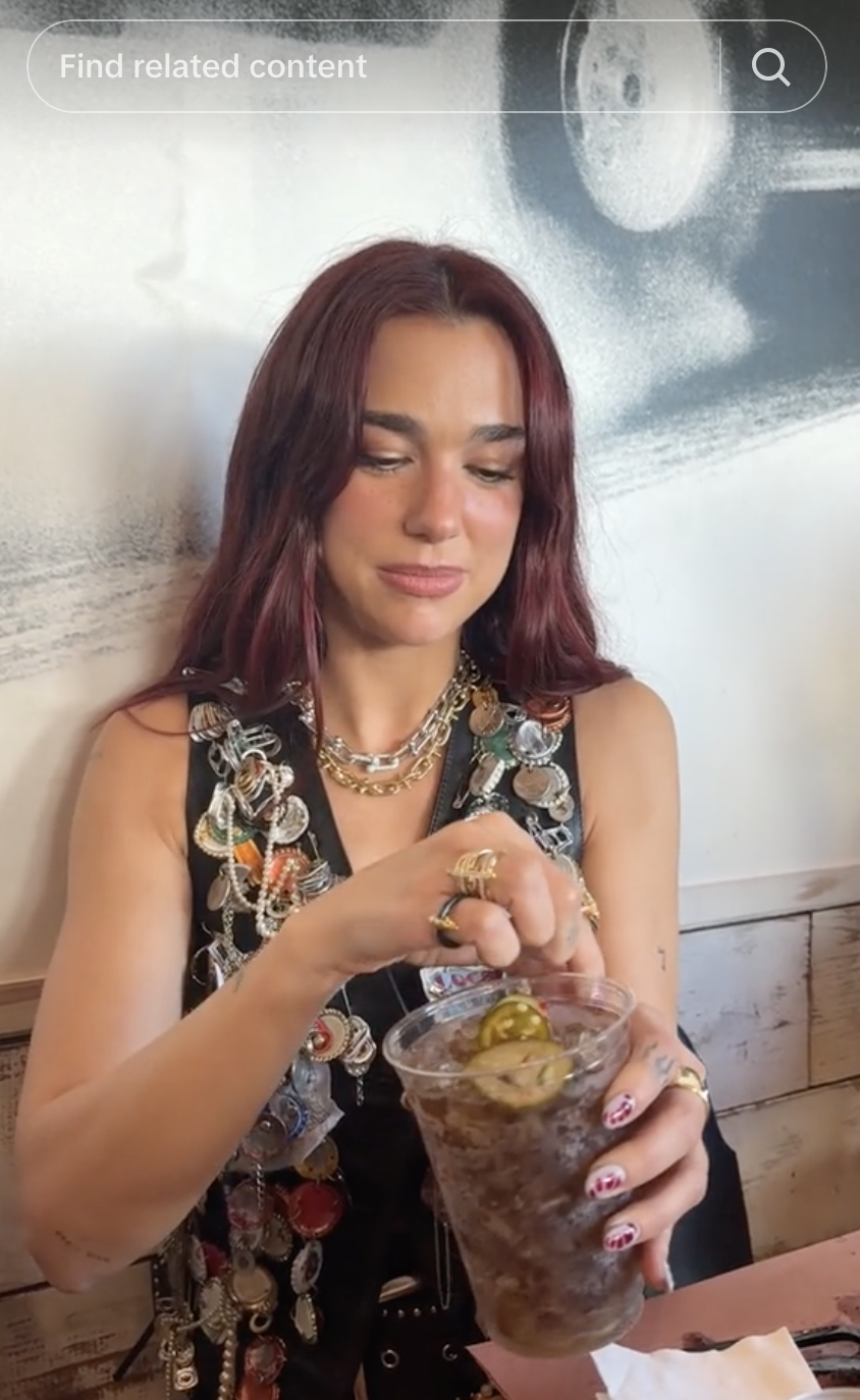 A person with long hair is stirring a drink with jalapeños, wearing a beaded vest and layered necklaces