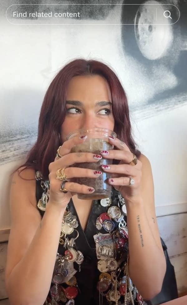 A person with shoulder-length hair drinks from a glass. They wear a vest adorned with various buttons and rings on their fingers