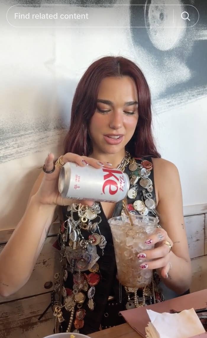 A person with long hair, wearing a vest with decorative buttons, is pouring a can of Diet Coke into a glass of ice at a table