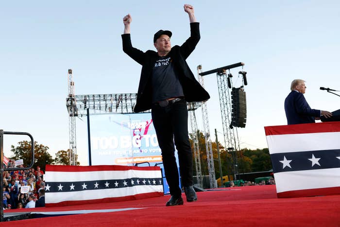 Elon Musk stands onstage with arms raised, wearing a black jacket and hat, at an outdoor event