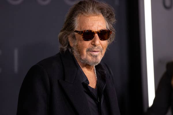 A person wearing sunglasses and a black suit jacket on a red carpet