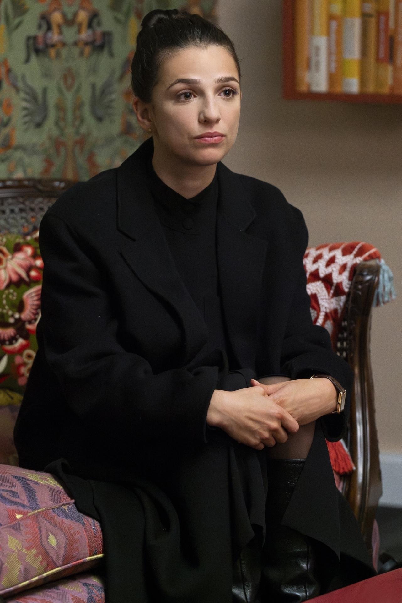A person sits on a patterned chair, wearing a black coat, with a neutral expression and hands clasped