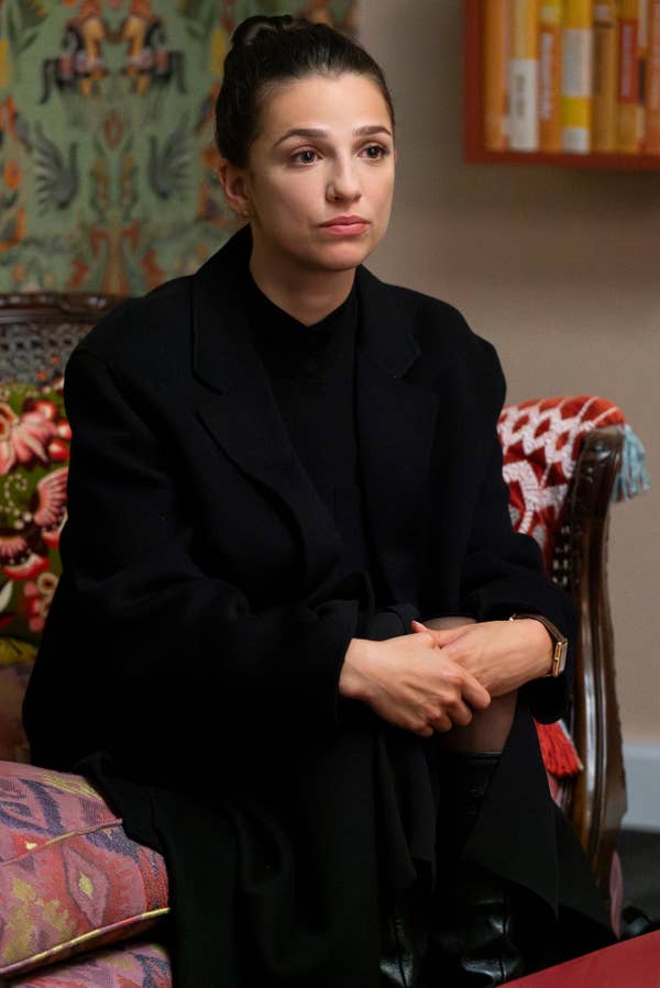 A person sits on a patterned chair, wearing a black coat, with a neutral expression and hands clasped
