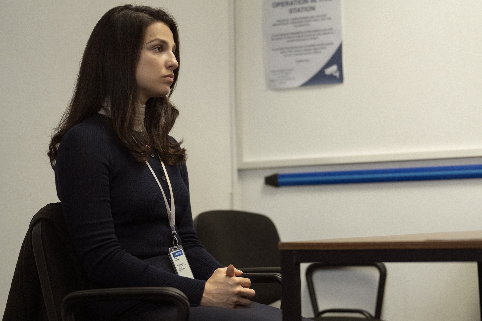 A woman sits in a chair at a desk in a waiting room, wearing a dark long-sleeve top and an ID badge. A poster is partially visible on the wall