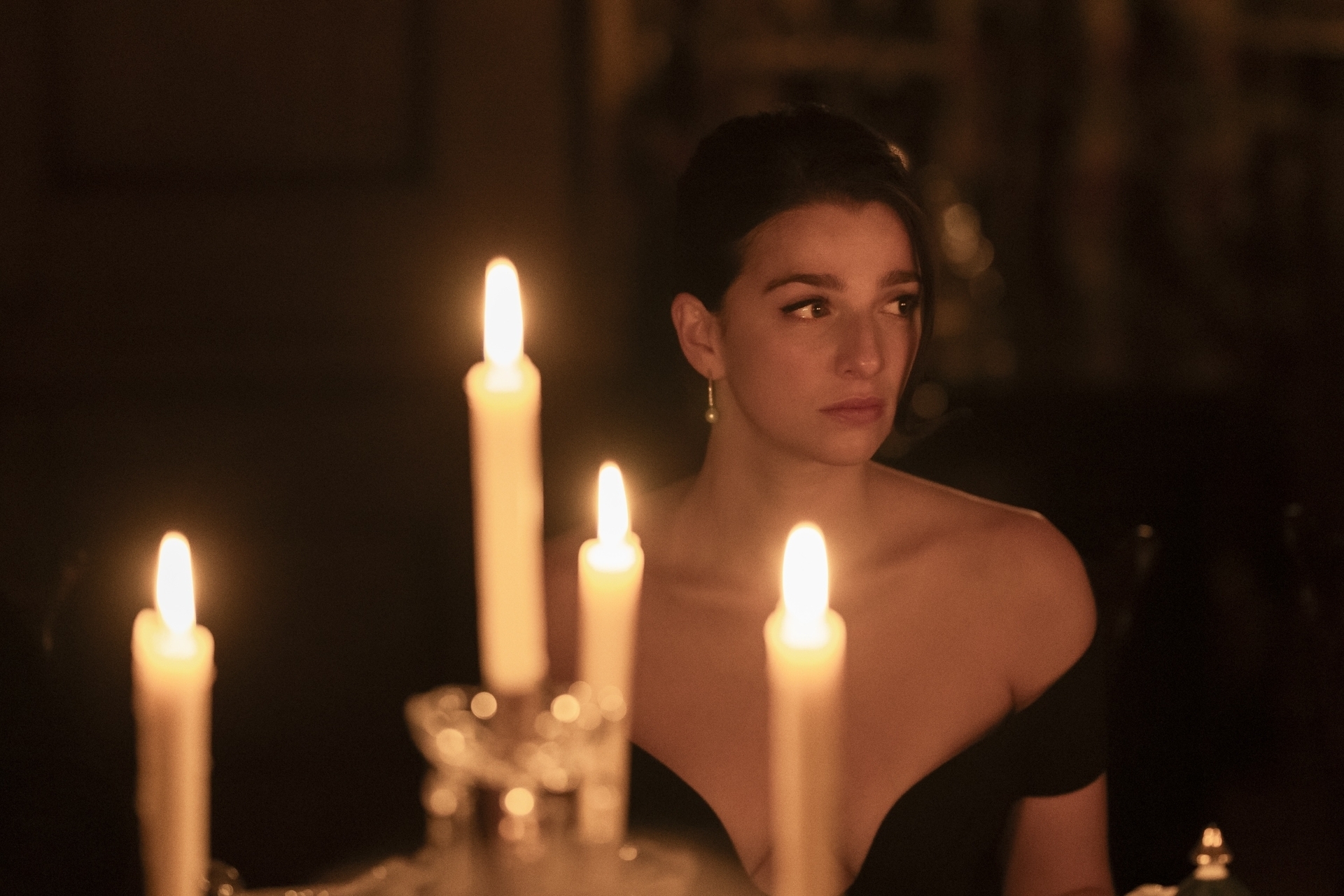I don't know who is in the image. A woman with short dark hair, wearing an off-the-shoulder dress, is sitting at a table with lit candles