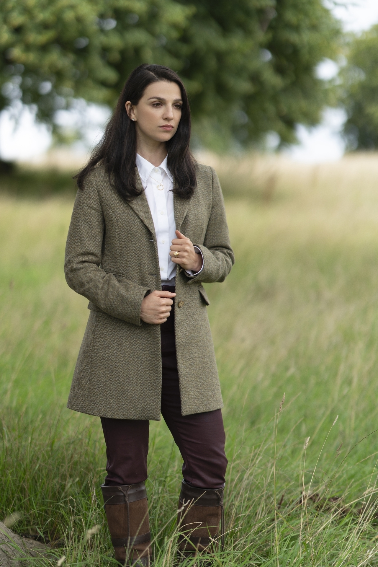 A person stands outdoors in a field wearing a tailored coat, white shirt, dark pants, and knee-high boots. They have long, dark hair