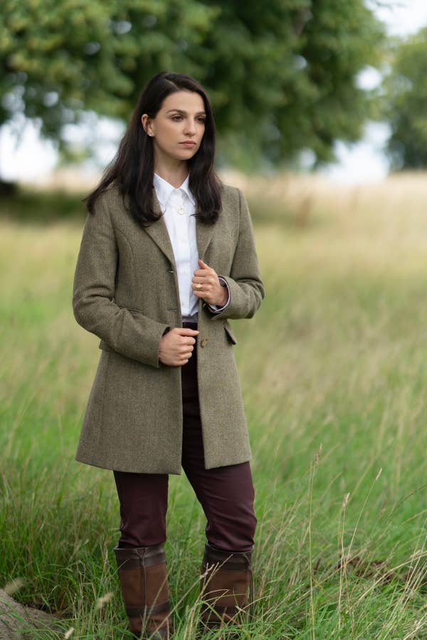 A person stands outdoors in a field wearing a tailored coat, white shirt, dark pants, and knee-high boots. They have long, dark hair