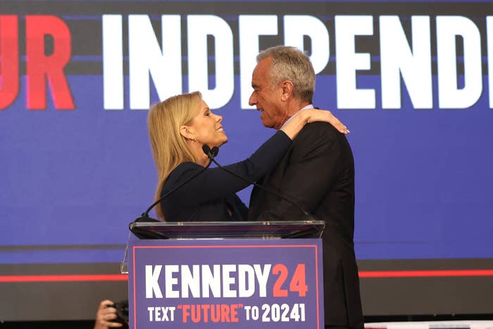 Two people embrace on stage at a Kennedy 2024 event. A sign reads "Text 'FUTURE' to 20241."
