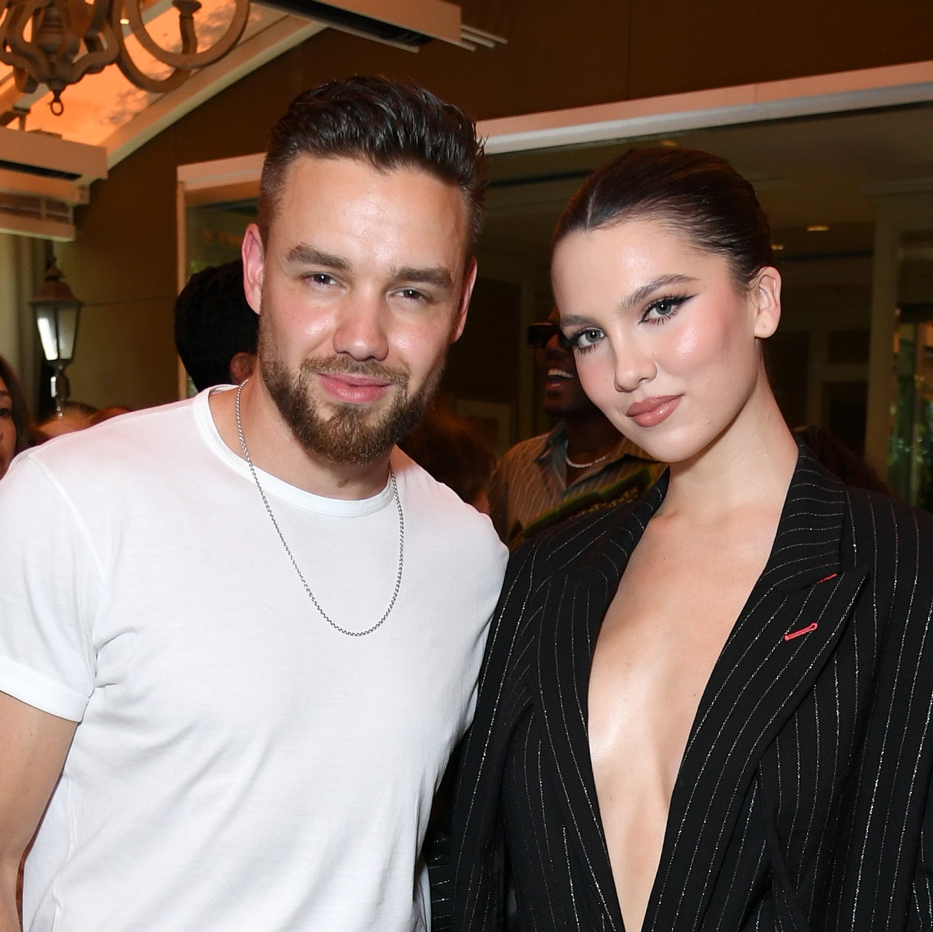 Liam Payne in a t-shirt and pants stands next to Maya Henry in a pinstripe suit with a deep neckline, posing at an indoor event