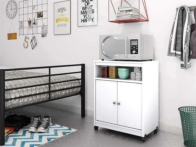 Microwave on a white storage cabinet in a bedroom with a bed, shelving, and decor items. Shopping article context. No people in the image