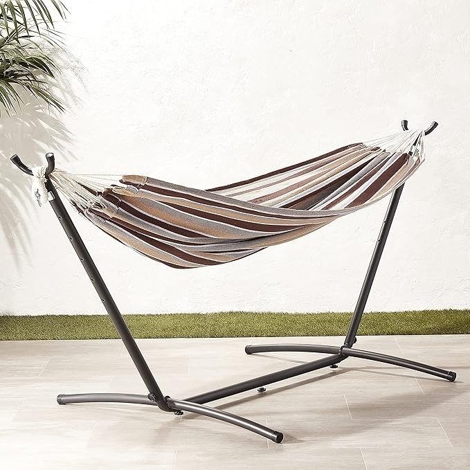 Hammock with a striped pattern on a sturdy metal stand, set indoors on a wooden floor with a potted plant nearby