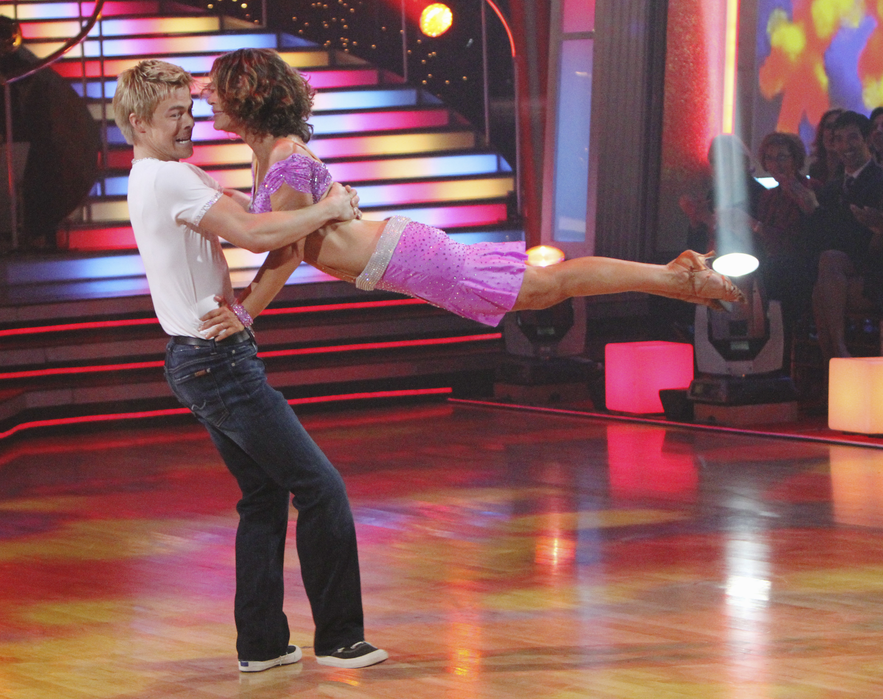 Jennifer Grey and Derek Hough perform a lively routine on a colorful stage; one lifts the other, displaying impressive balance and form