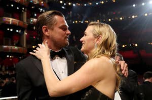 Leonardo DiCaprio and Kate Winslet embrace and smile at each other during an event in a large auditorium. DiCaprio wears a black tuxedo, Winslet wears a formal dress