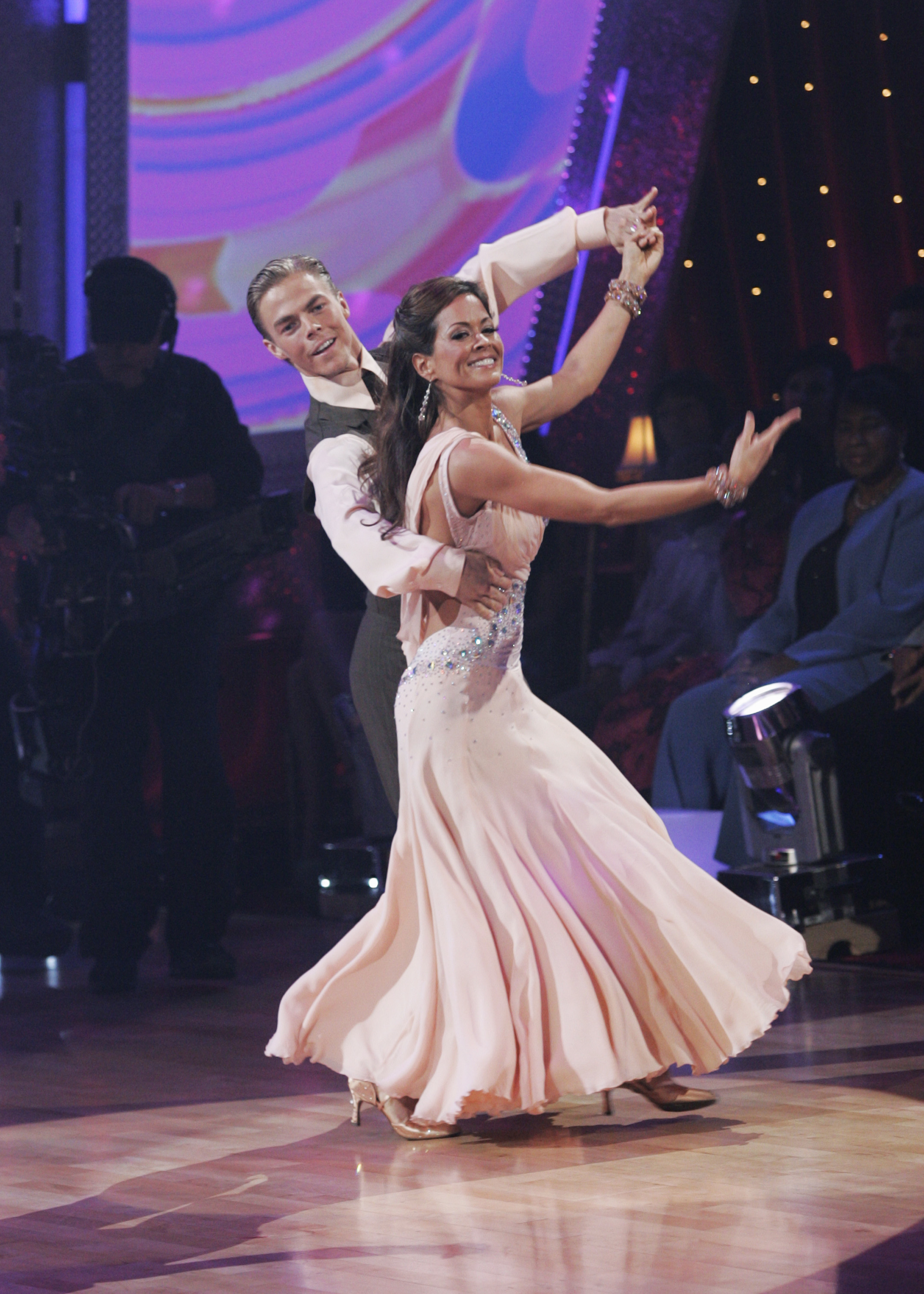 Two dancers in elegant ballroom attire perform energetically on a stage with an audience in the background