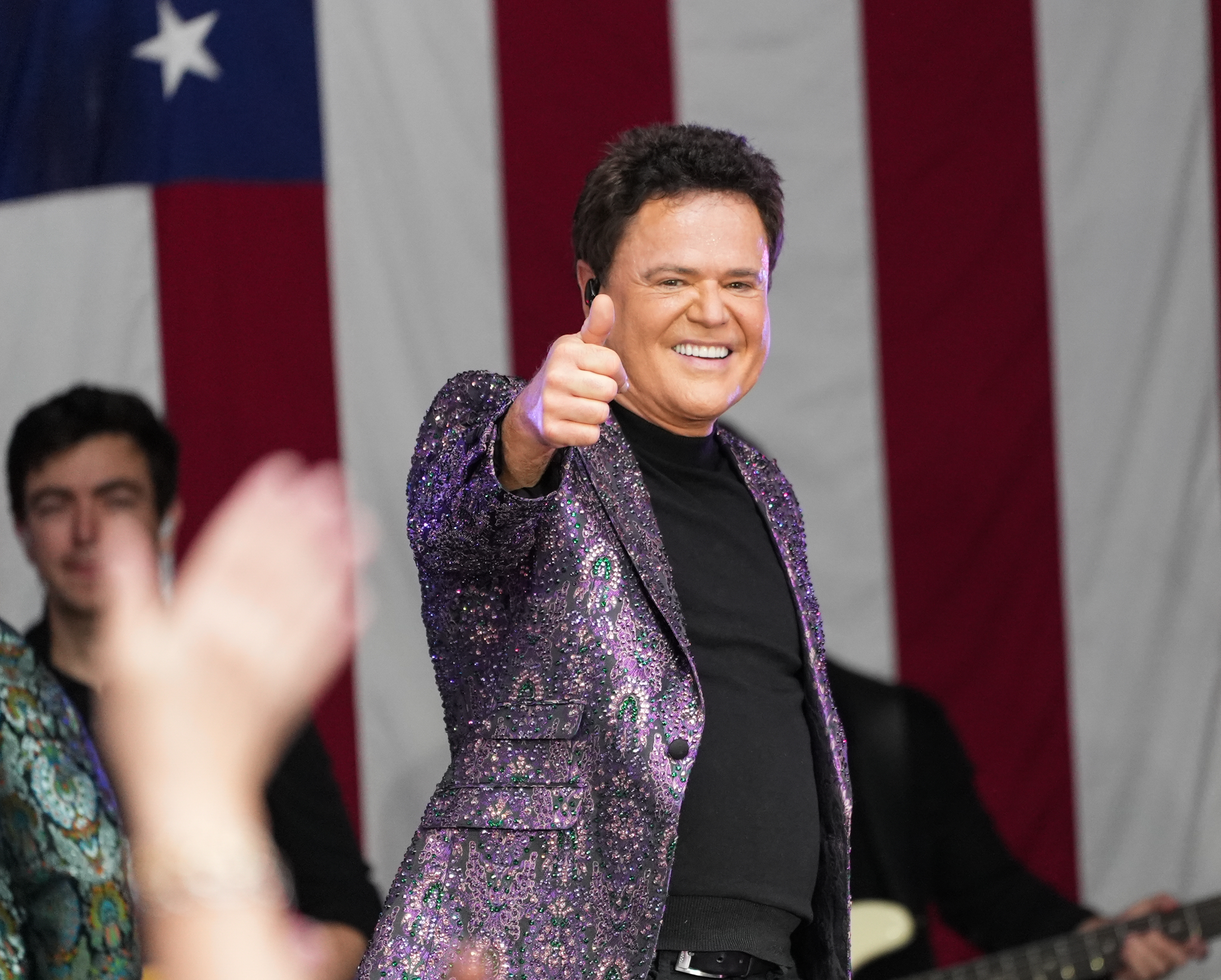 A person in a sparkling suit gives a thumbs up on stage, with a U.S. flag in the background