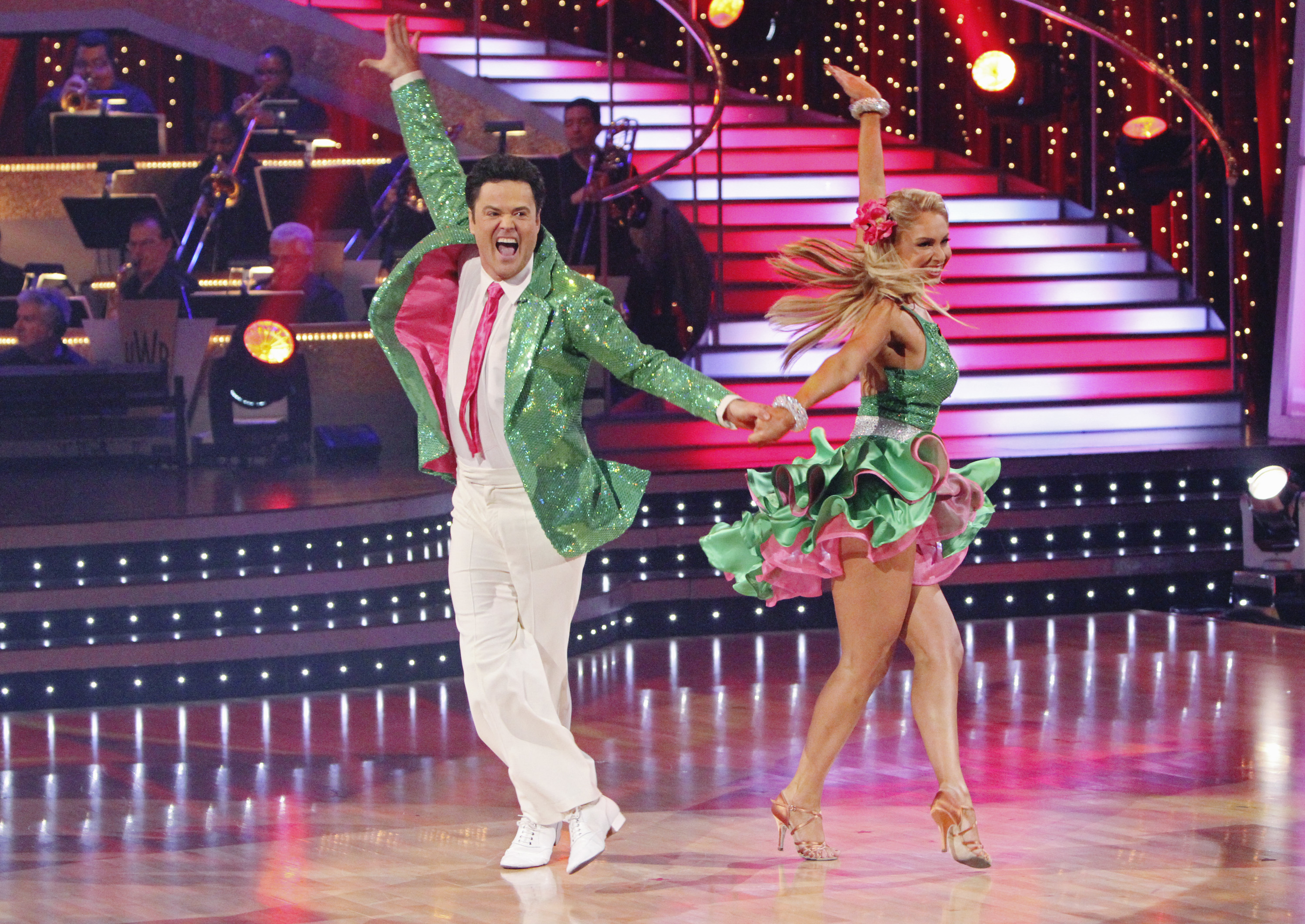 Donny Osmond and Kym Johnson perform energetically with coordinated outfits on a lit stage, surrounded by vibrant lighting and a band in the background