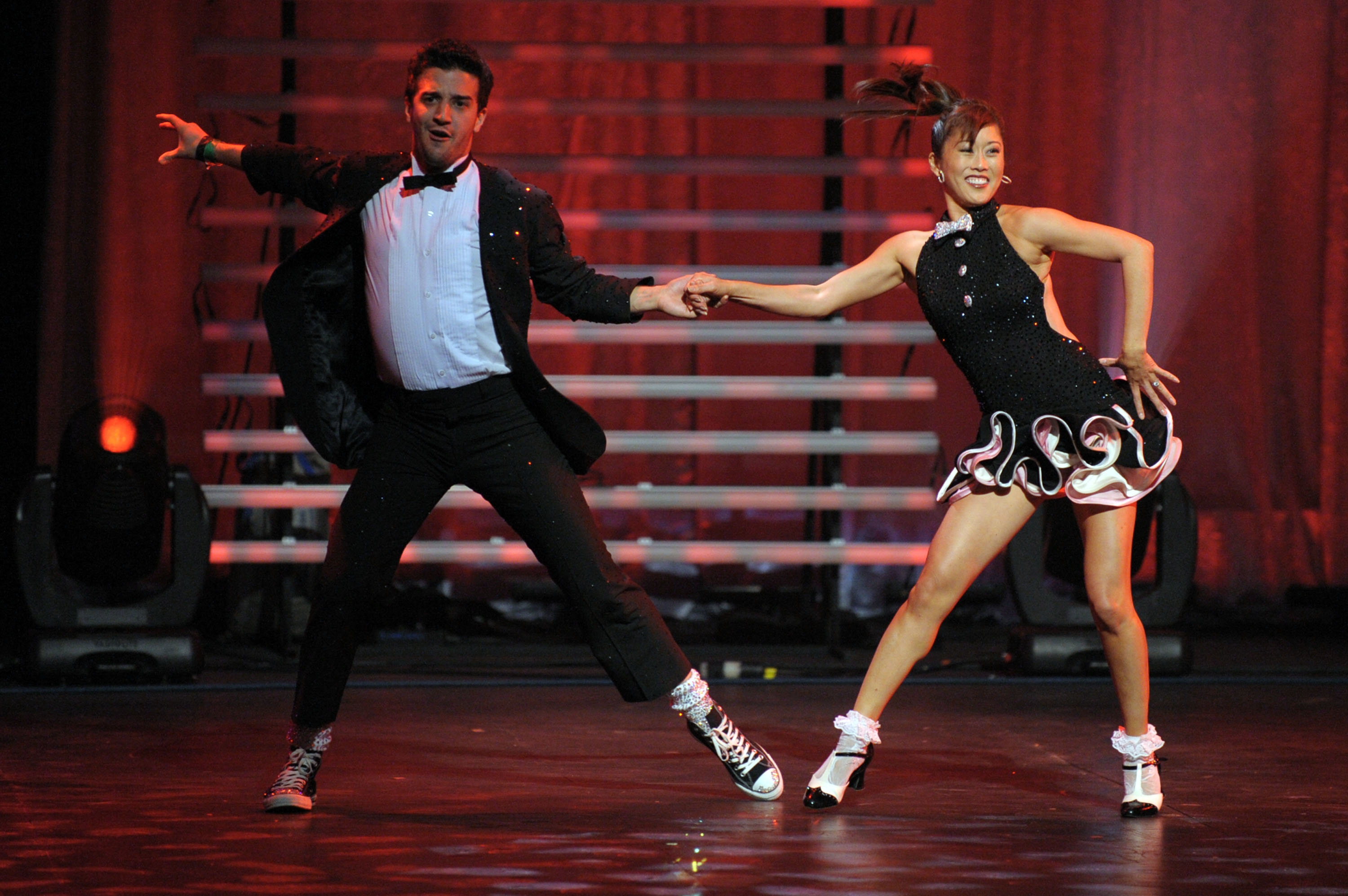 Dancer Mark Ballas and figure skater Kristi Yamaguchi are dancing energetically on stage in formal attire, with one wearing a suit and the other in a short dress