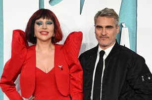 Two people on a red carpet. One wears a dramatic gown with structured shoulders, the other is in a suit with sneakers. Names not provided