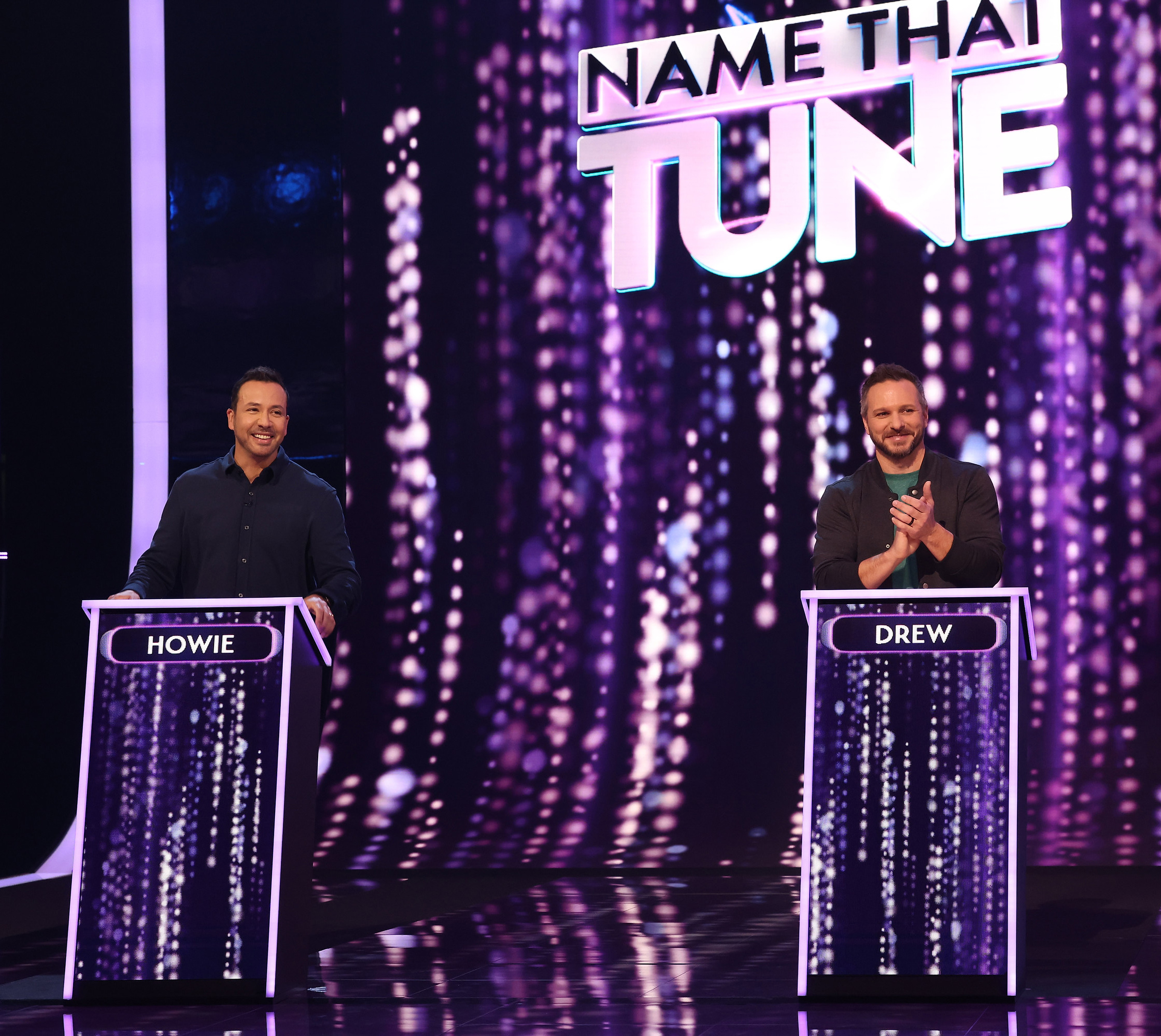 Two contestants, Howie and Drew, stand at podiums on the game show &quot;Name That Tune.&quot; They&#x27;re smiling under the stage lights