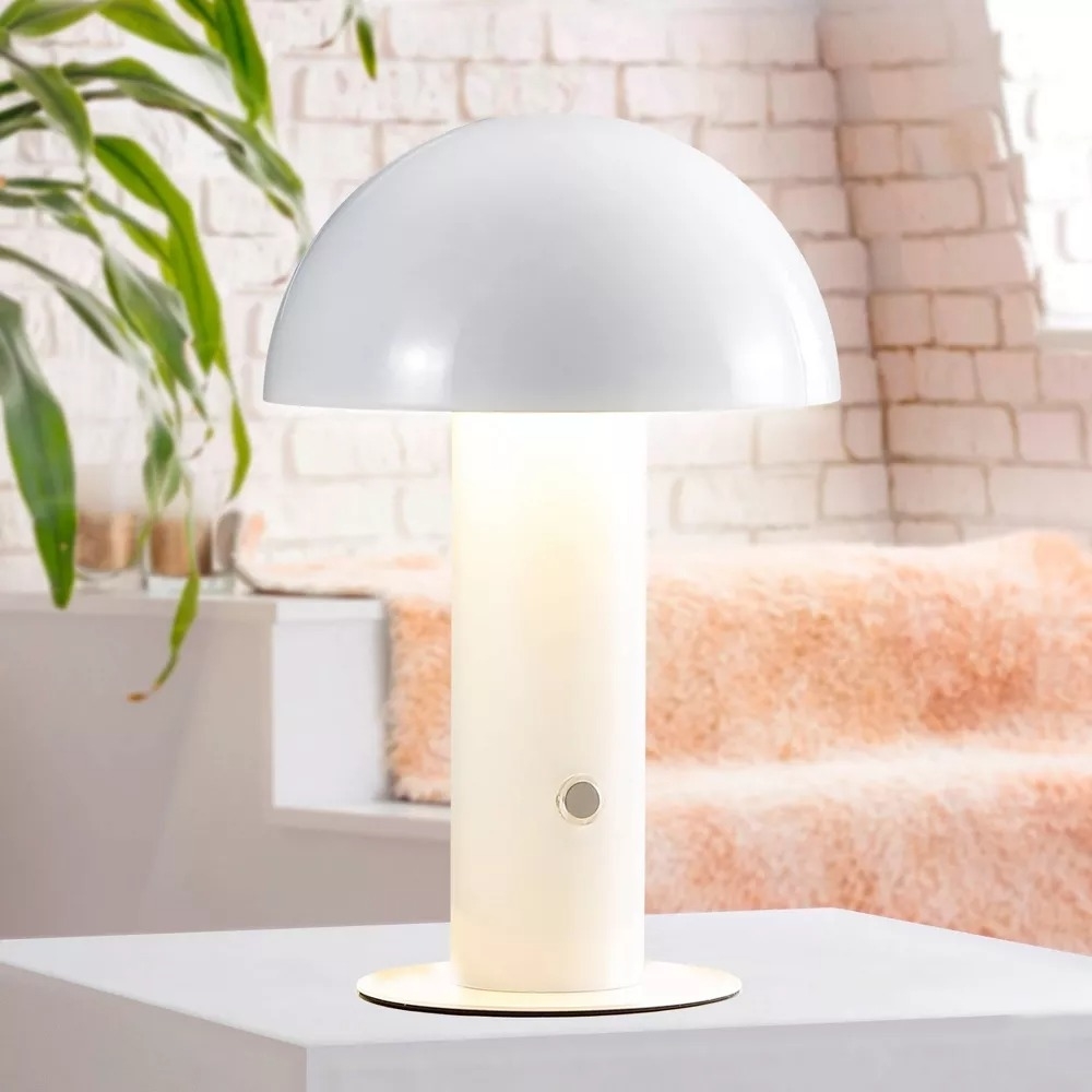 Modern table lamp with a dome-shaped shade on a cylindrical base, placed on a white surface in a cozy room with plants and soft fabrics