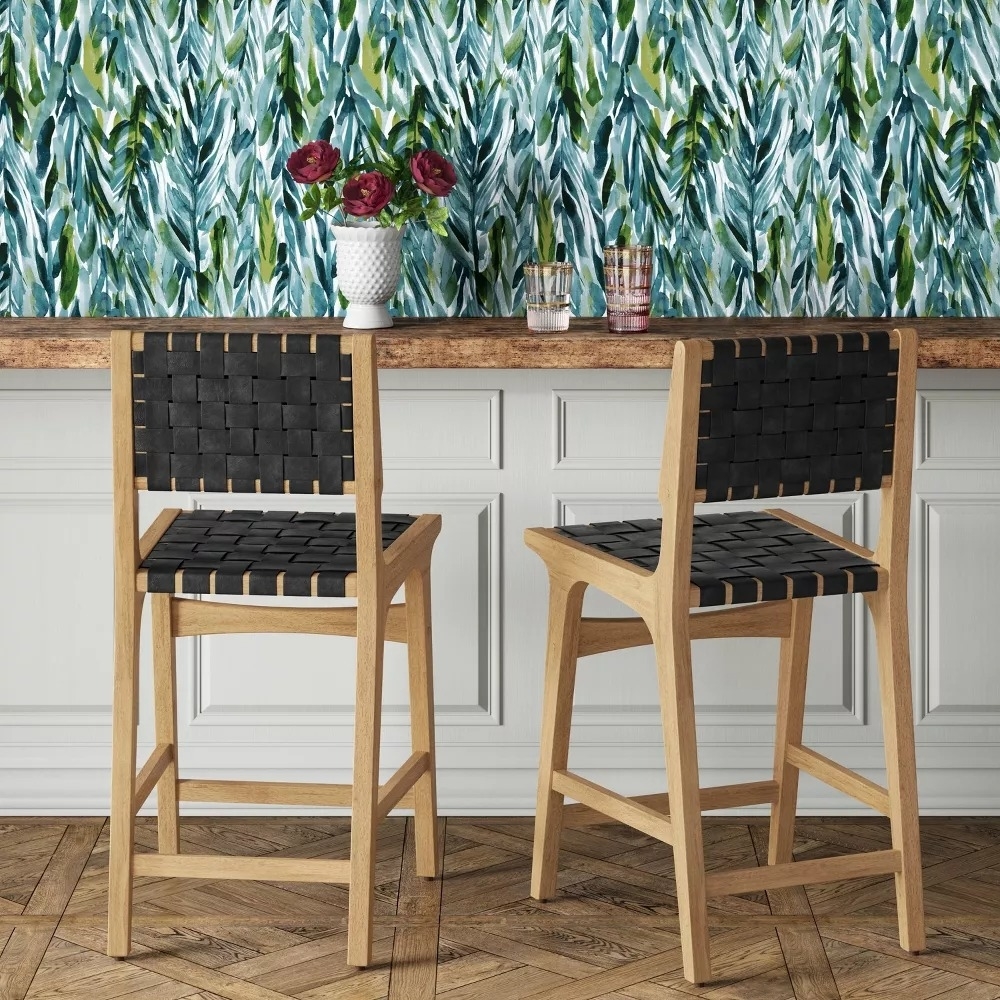 Two woven black leather barstools at a wooden counter, with a decorative plant and glasses against a leafy patterned wallpaper backdrop