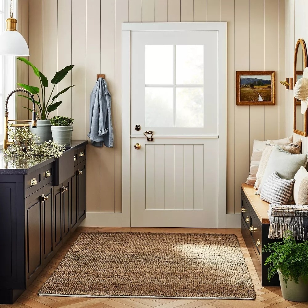 A cozy entryway features a white door, black cabinets with plants, a bench with cushions, and a woven rug, creating a warm and inviting space