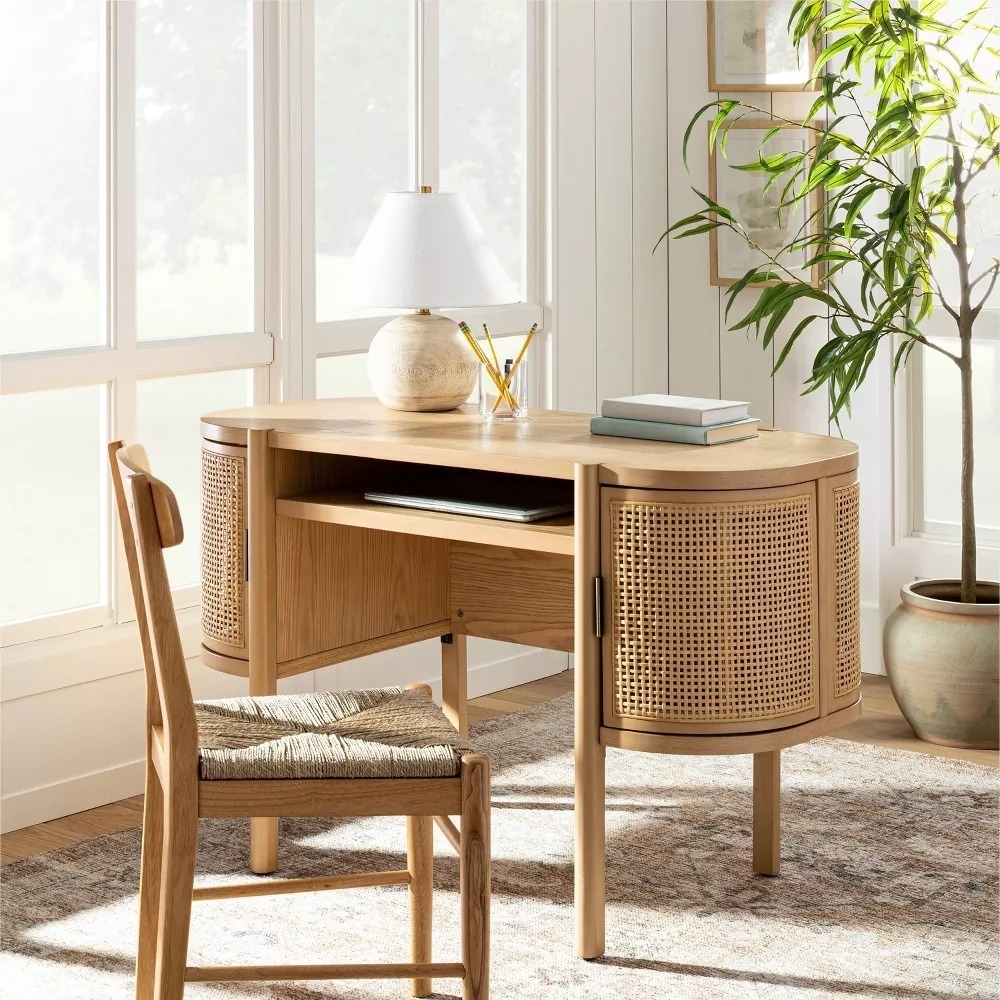 Modern wooden desk with a round lamp, books, and a plant nearby, situated by large windows in a bright room