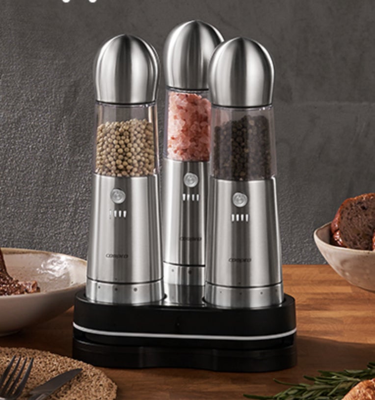 Electric salt, pepper, and spice grinders on a wooden table, with food partially visible nearby