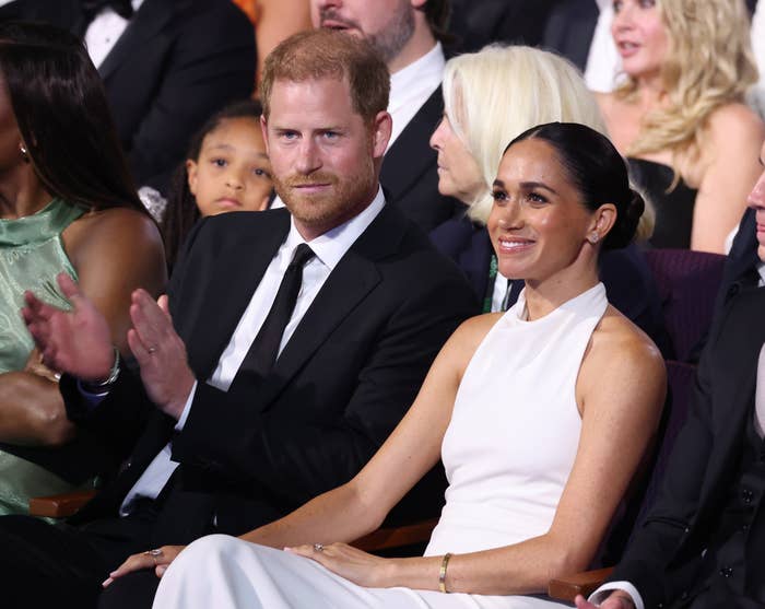 Prince Harry and Megan sit together in formal wear at an event, surrounded by others
