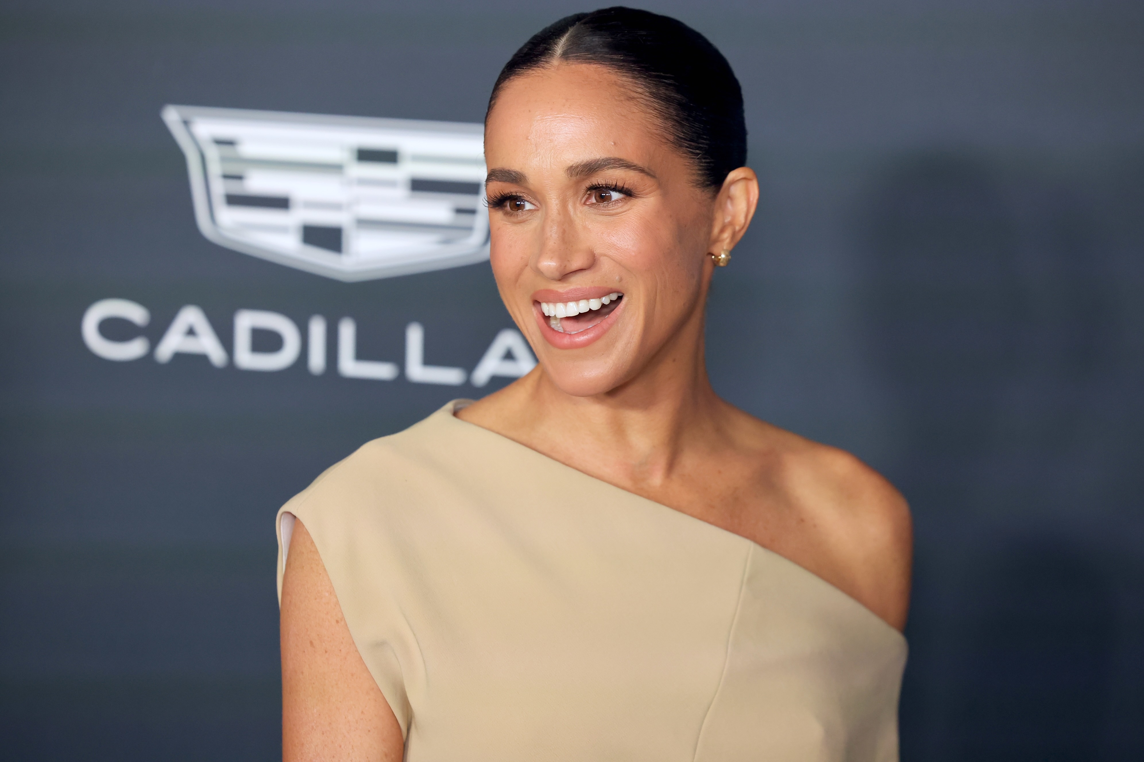 Megan in elegant one-shoulder attire smiles at an event, with a Cadillac logo in the background