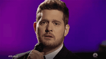Michael Buble holds a microphone on stage and winks. The background is purple