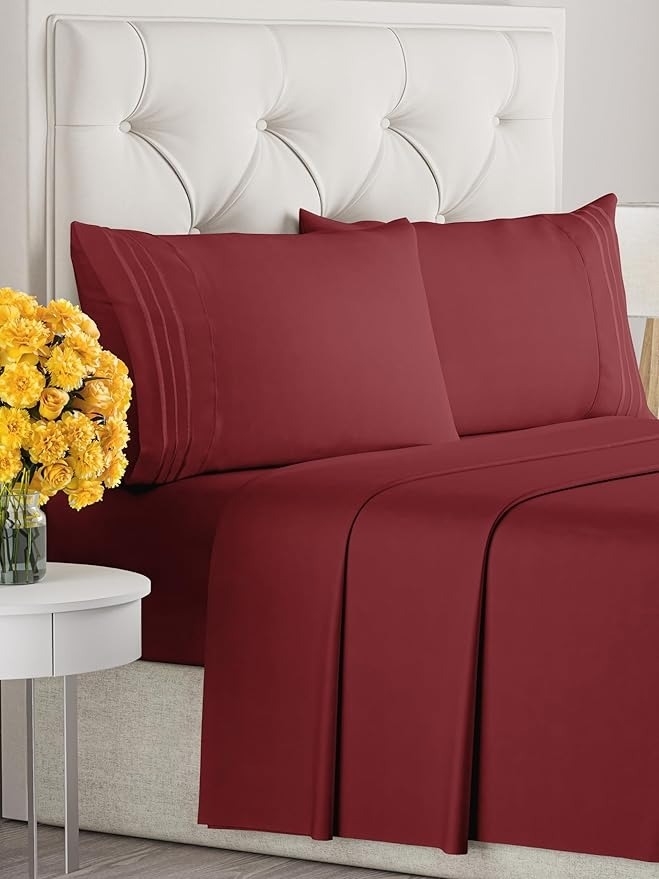 Luxurious red bed sheets and pillowcases on a tufted headboard with yellow flowers on a bedside table