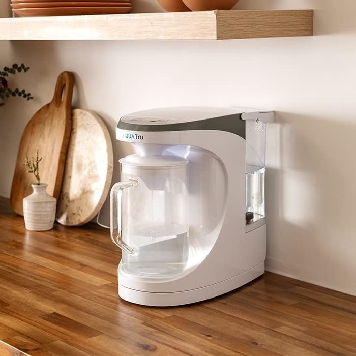 Countertop water purifier with a pitcher on a wooden kitchen counter, surrounded by decorative cutting boards