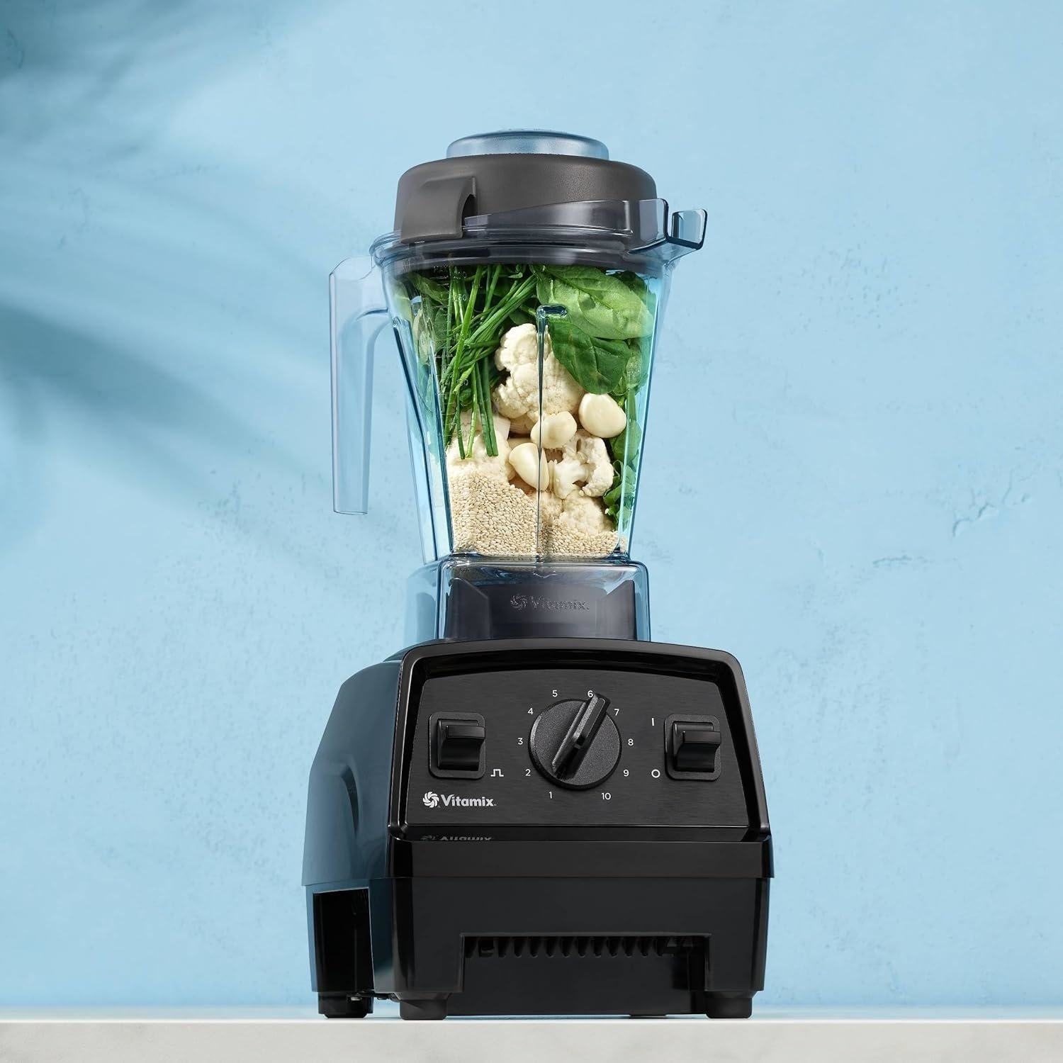 Vitamix blender filled with ingredients including cauliflower, quinoa, and greens, against a plain background