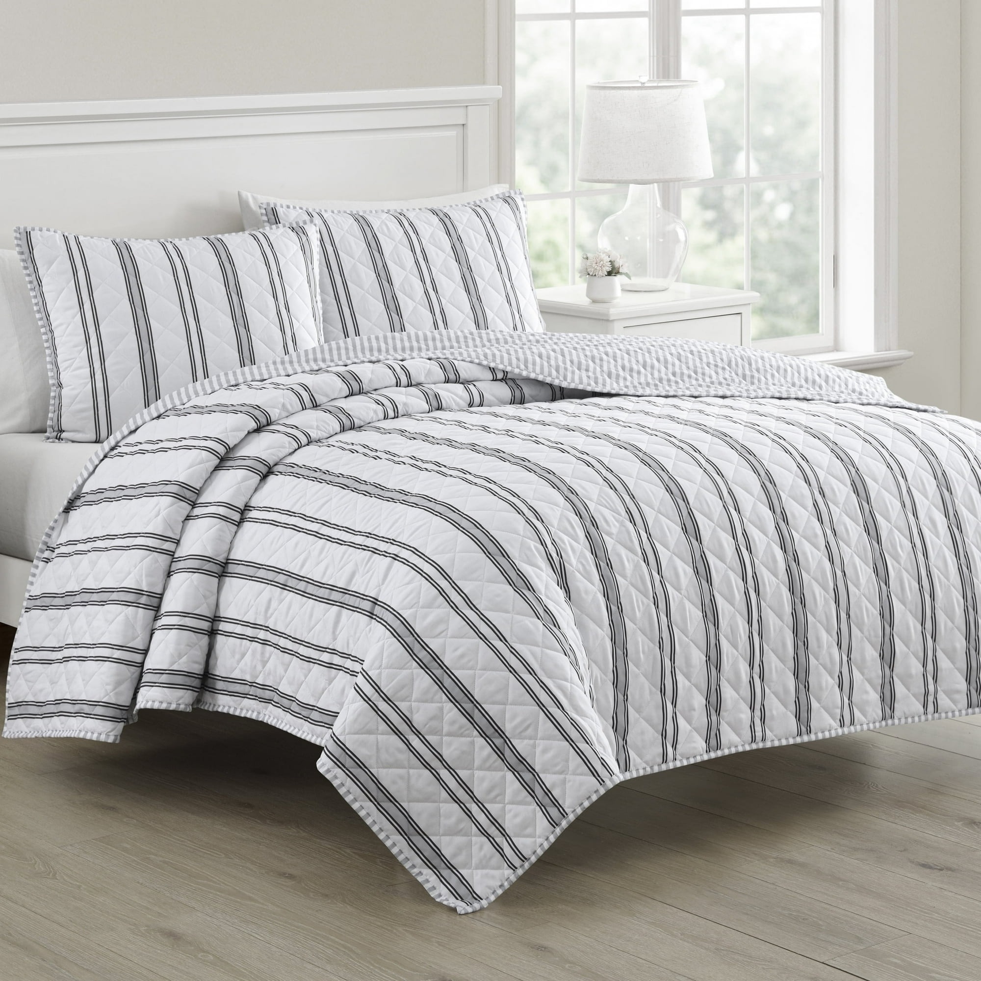 Striped quilt and pillow set on a neatly made bed in a bright room, featuring large windows and a bedside lamp