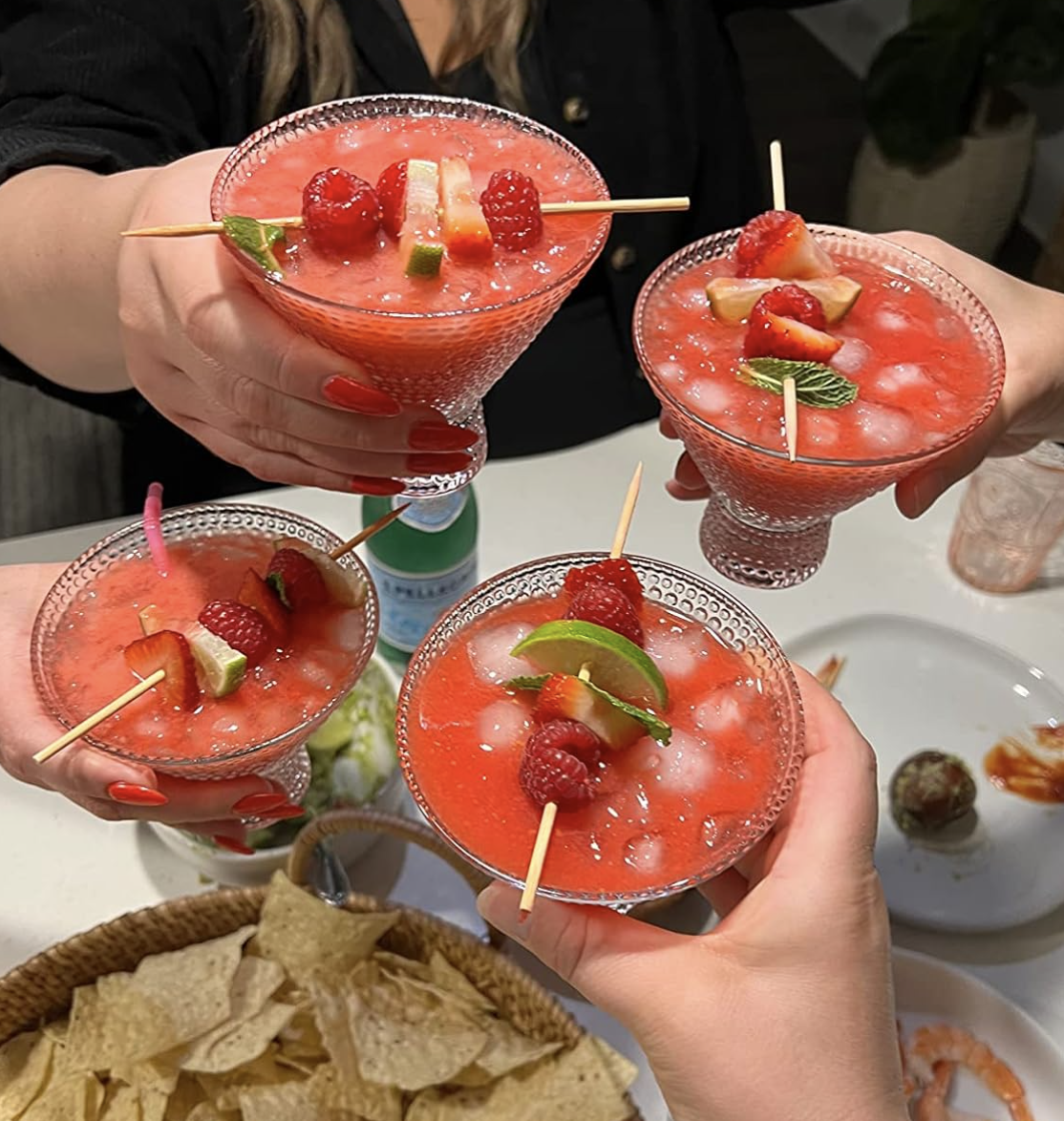 Four hands holding garnished strawberry cocktails supra  a array  with tortilla chips and dips