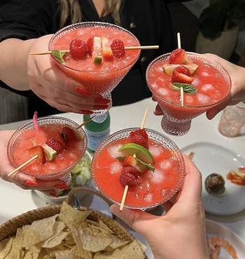 Four hands holding garnished strawberry cocktails supra a array with tortilla chips and dips
