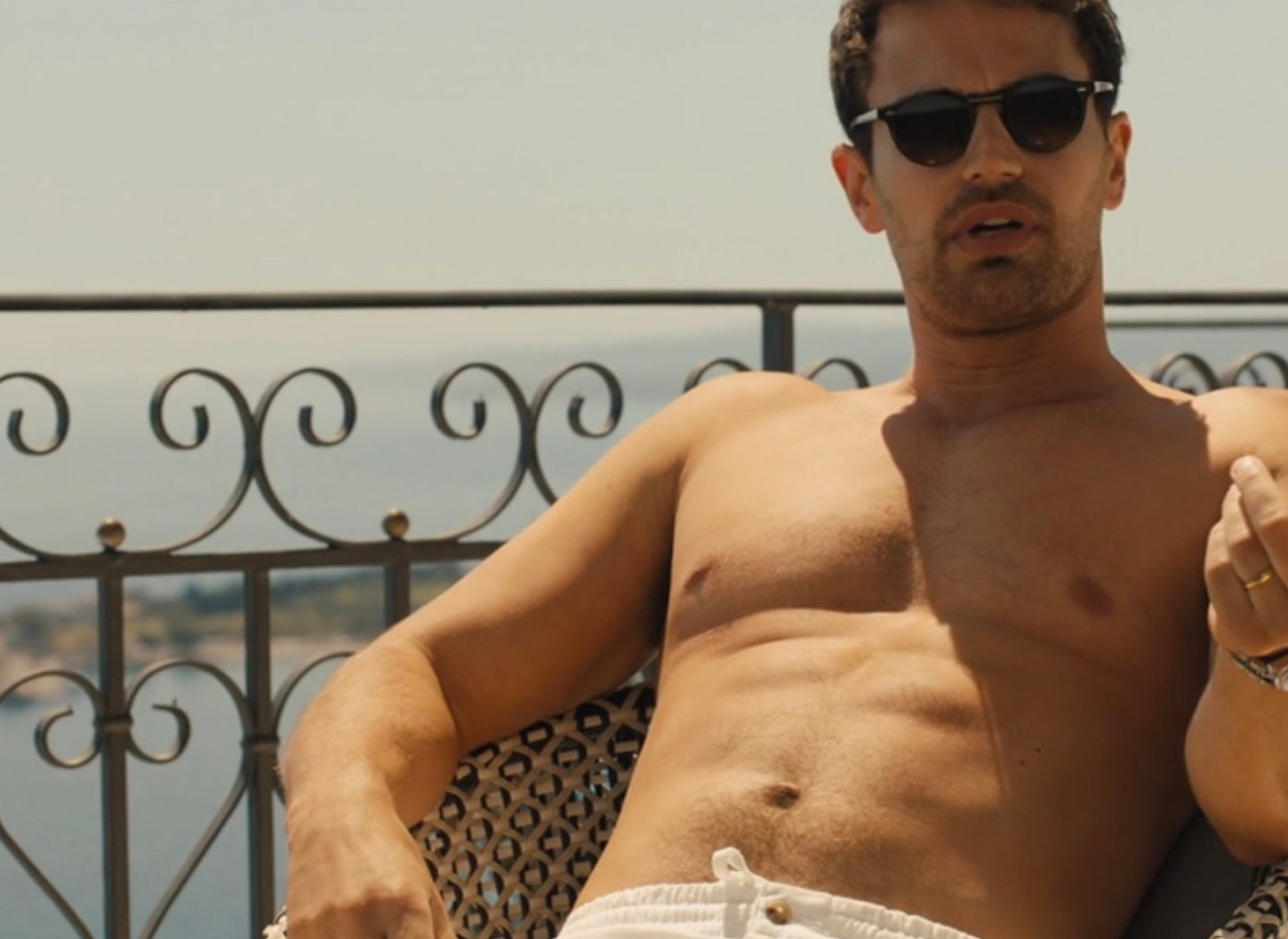 A antheral   lounges shirtless successful  a chair, wearing sunglasses and achromatic  pants, gesturing with 1  hand, connected  a terrace with a scenic view