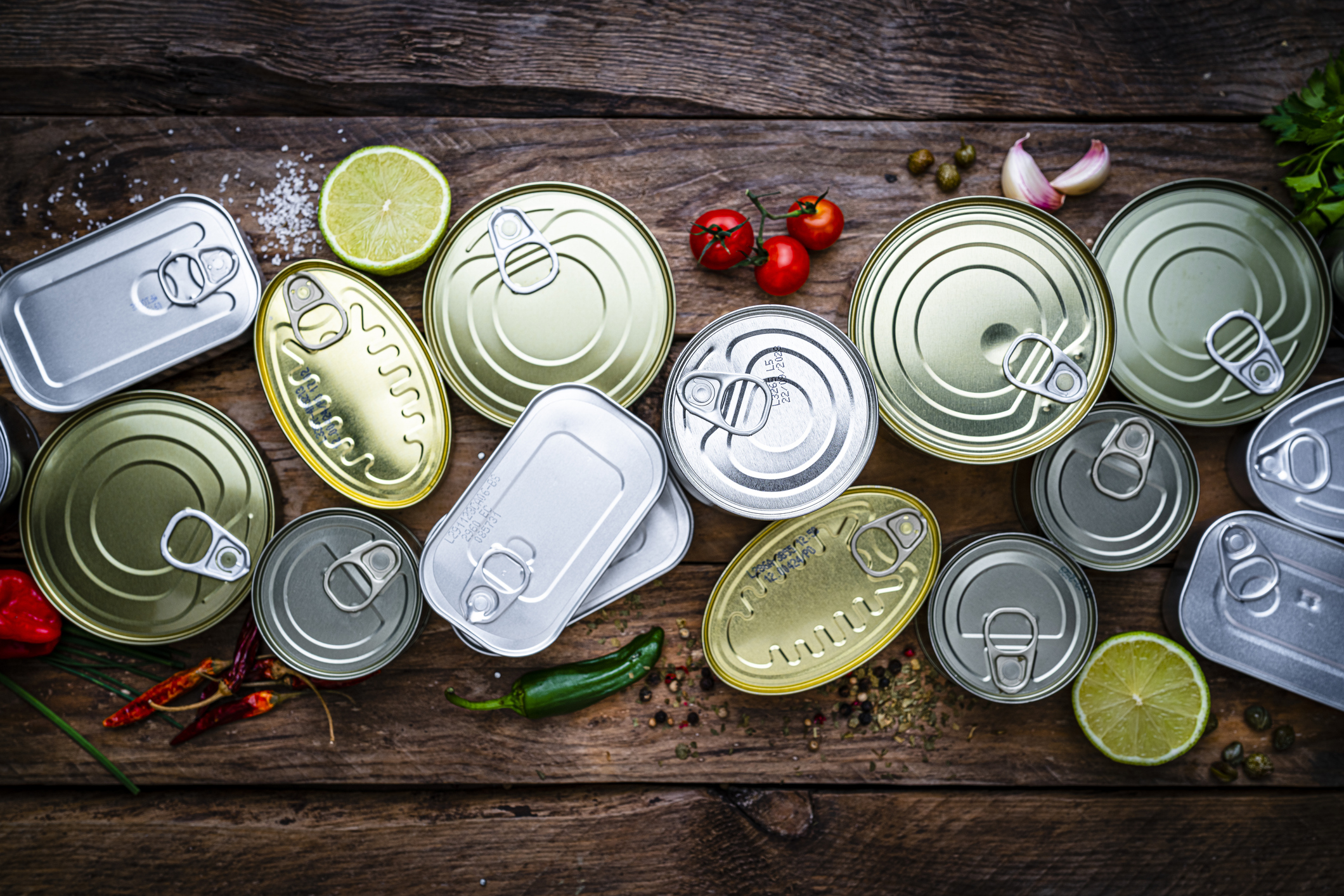 Various unopened canned goods, fresh lime halves, tomatoes, garlic cloves, and chili peppers on a wooden surface