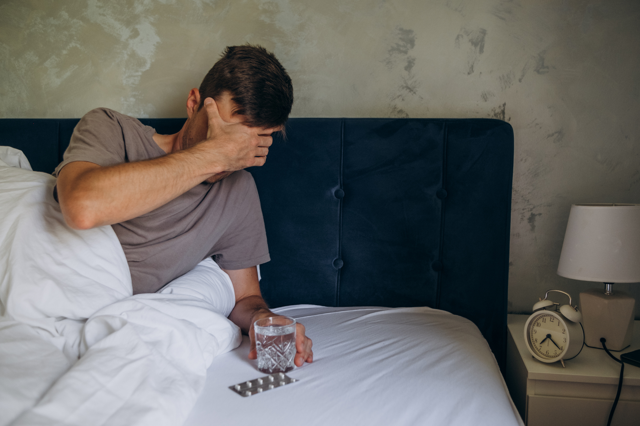 A person sits in bed with a headache, holding a glass of water and a blister pack of pills on the bed. An alarm clock sits on the bedside table